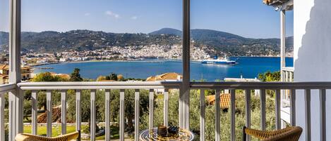 A balcony with wicker chairs and a small table, overlooking a coastal town and a body of water.