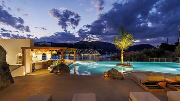 Área de piscina com espreguiçadeiras, bar e vista para a paisagem urbana ao entardecer.