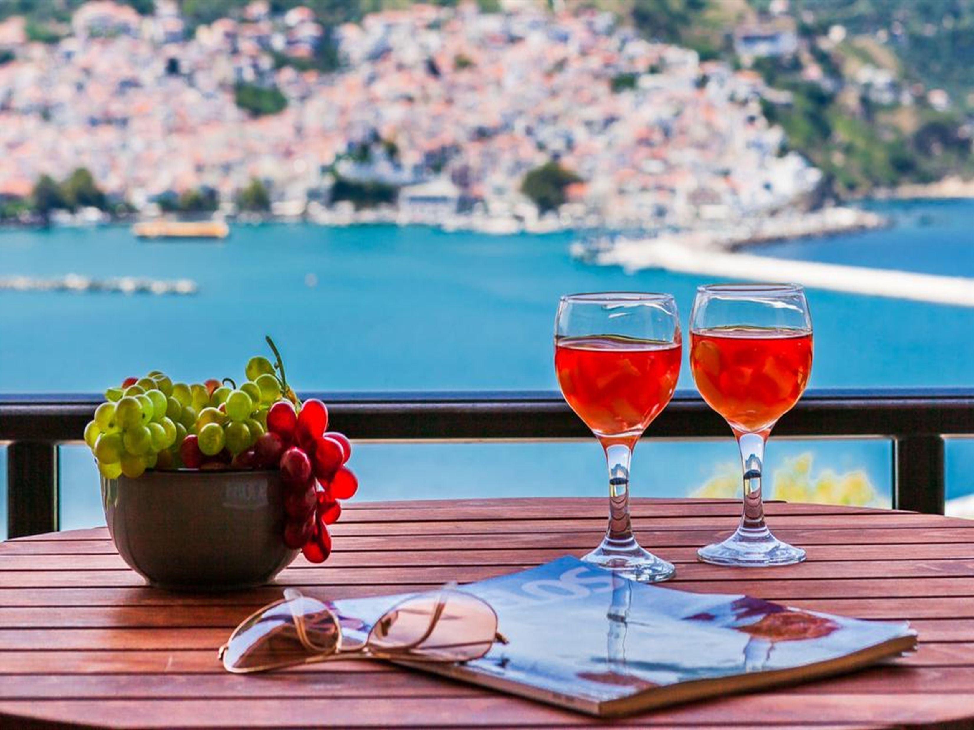 A wooden table with a bowl of fruit, two wine glasses, and a pair of sunglasses.
