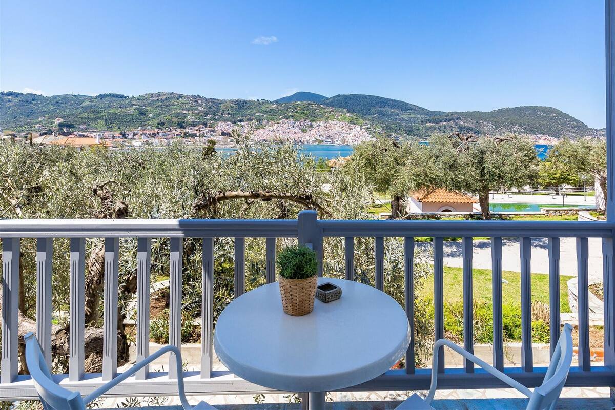 Un balcon avec une table et des chaises, donnant sur un paysage côtier parsemé d’oliviers et sur une ville située au loin.