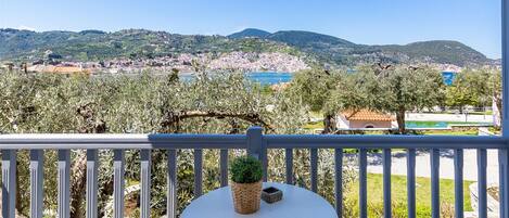 A balcony with a table and chairs, overlooking a coastal landscape with olive trees and a distant town.