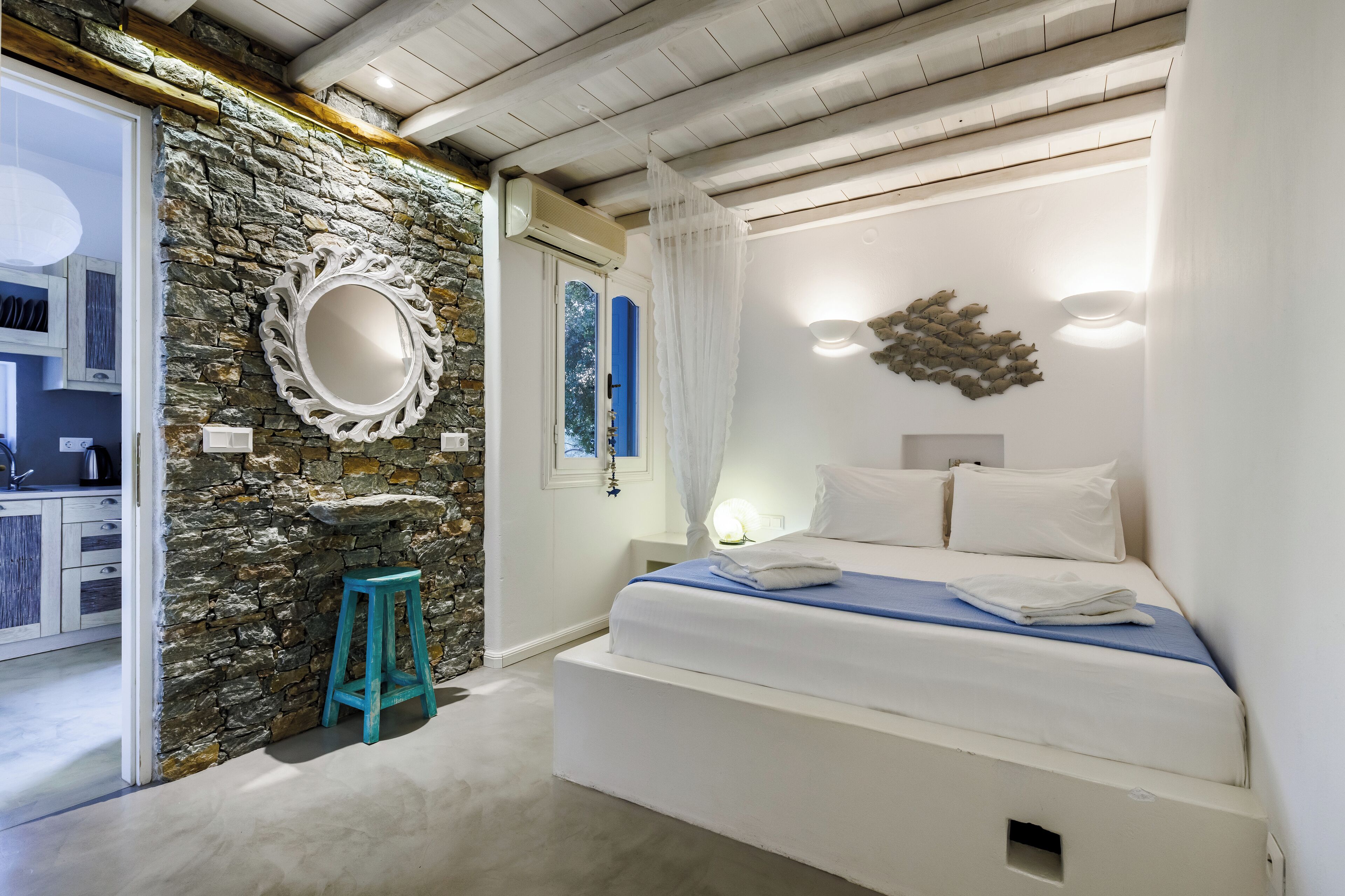 A bedroom with a stone wall, a bed with white bedding, a blue stool, and a mirror.
