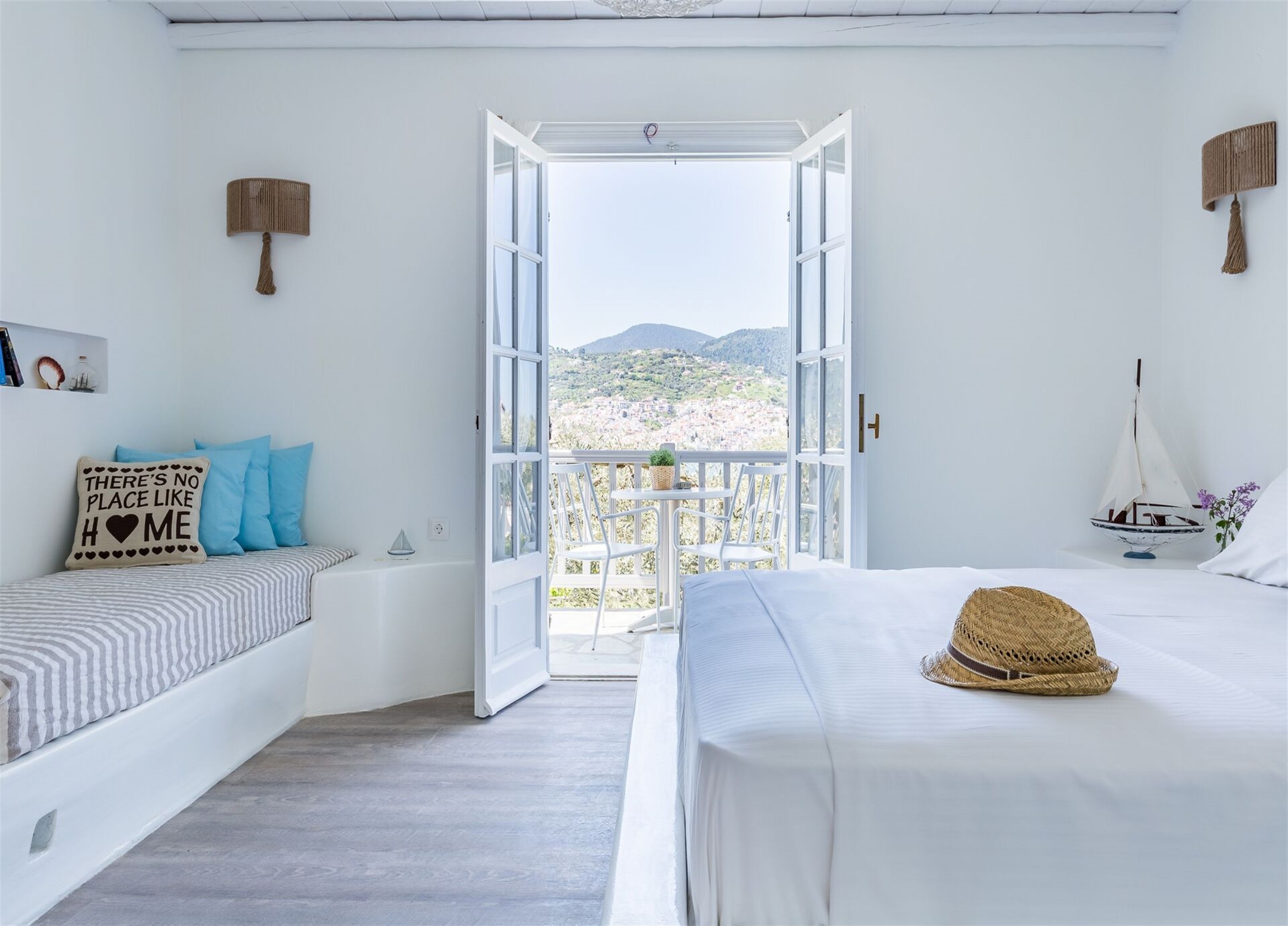 A bedroom with a bed, a bedside table, a chair, a small table with a plant, and a view of a town and mountains through an open door.