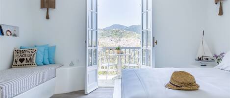 A bedroom with a bed, a bedside table, a chair, a small table with a plant, and a view of a town and mountains through an open door.