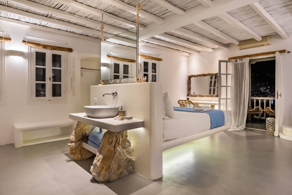 Une salle de bain moderne avec un lavabo en pierre, un banc et un lit offrant une vue sur la ville la nuit.