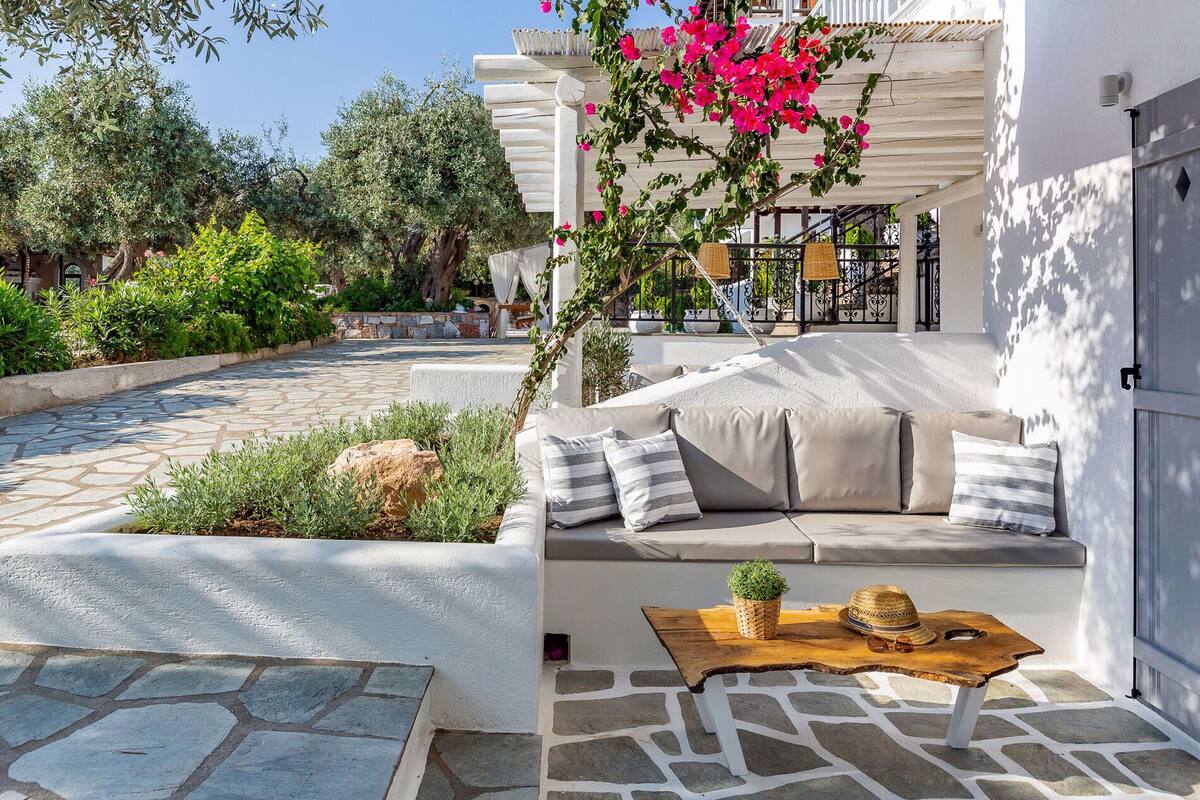 Un espace extérieur ensoleillé avec des sièges, une terrasse carrelée en pierre, une table en bois agrémentée d’un chapeau et d’une plante, et un mur blanc avec un balcon.