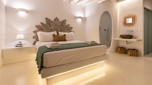 A modern bedroom with a large bed, bedside lamps, a decorative leaf headboard, a built-in shelf, and a small bathroom with a mirror and sink. - Evlalia Studios & Villas (Skopelos)