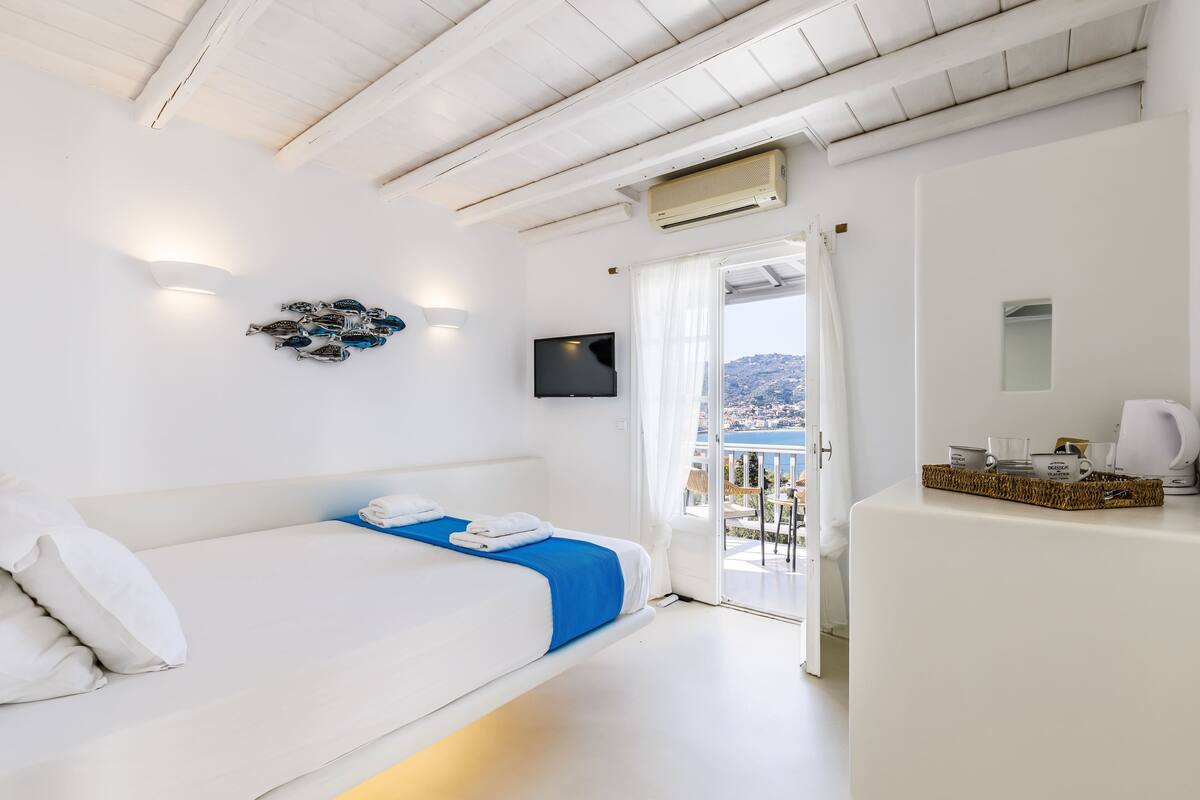 Une chambre d’hôtel avec un lit, une télévision à écran plat, une vue sur la mer et un balcon équipé d’une table et de chaises.