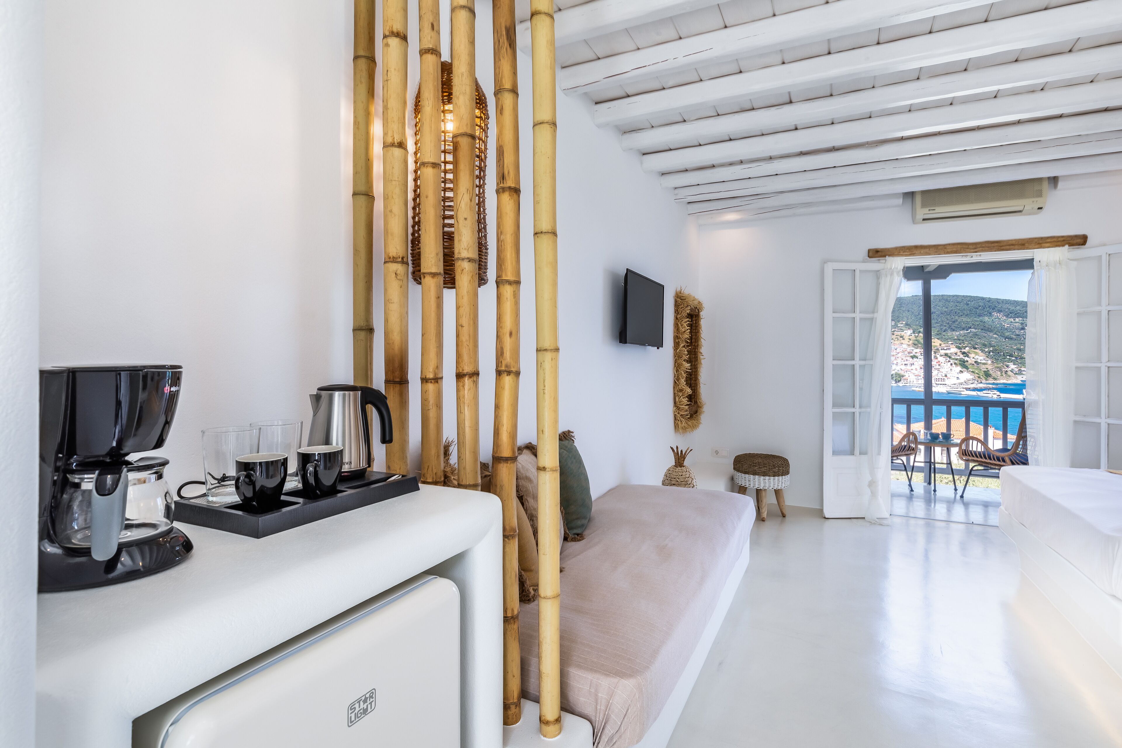 A modern hotel room with a white interior, a bed, a sofa, a coffee station, and a view of the sea through an open door.