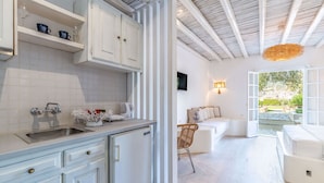 A modern kitchen with white cabinets, a built-in sink, and a dining area with a white sofa and a wicker chair. - Evlalia Studios & Villas (Skopelos)