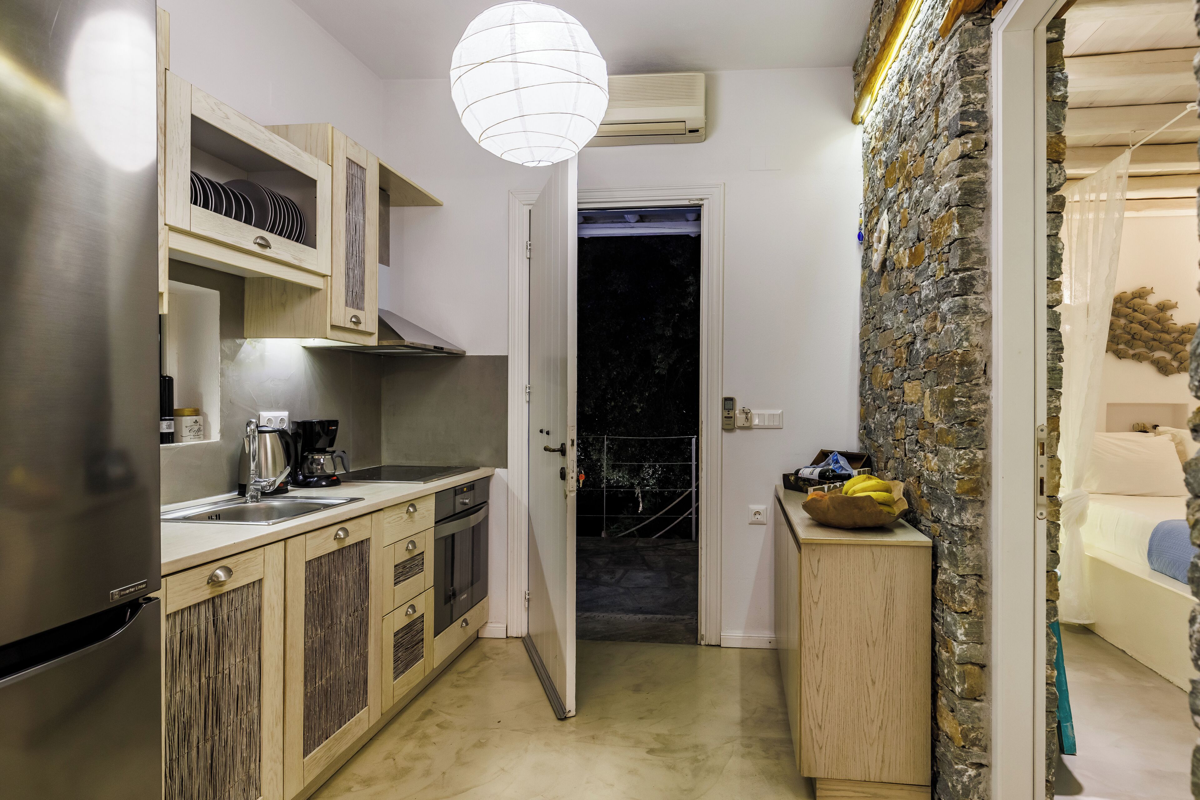 A compact kitchen with wooden cabinets, a stainless steel refrigerator, and a coffee maker. A stone wall and a bed with a blue blanket are visible in the adjacent room.