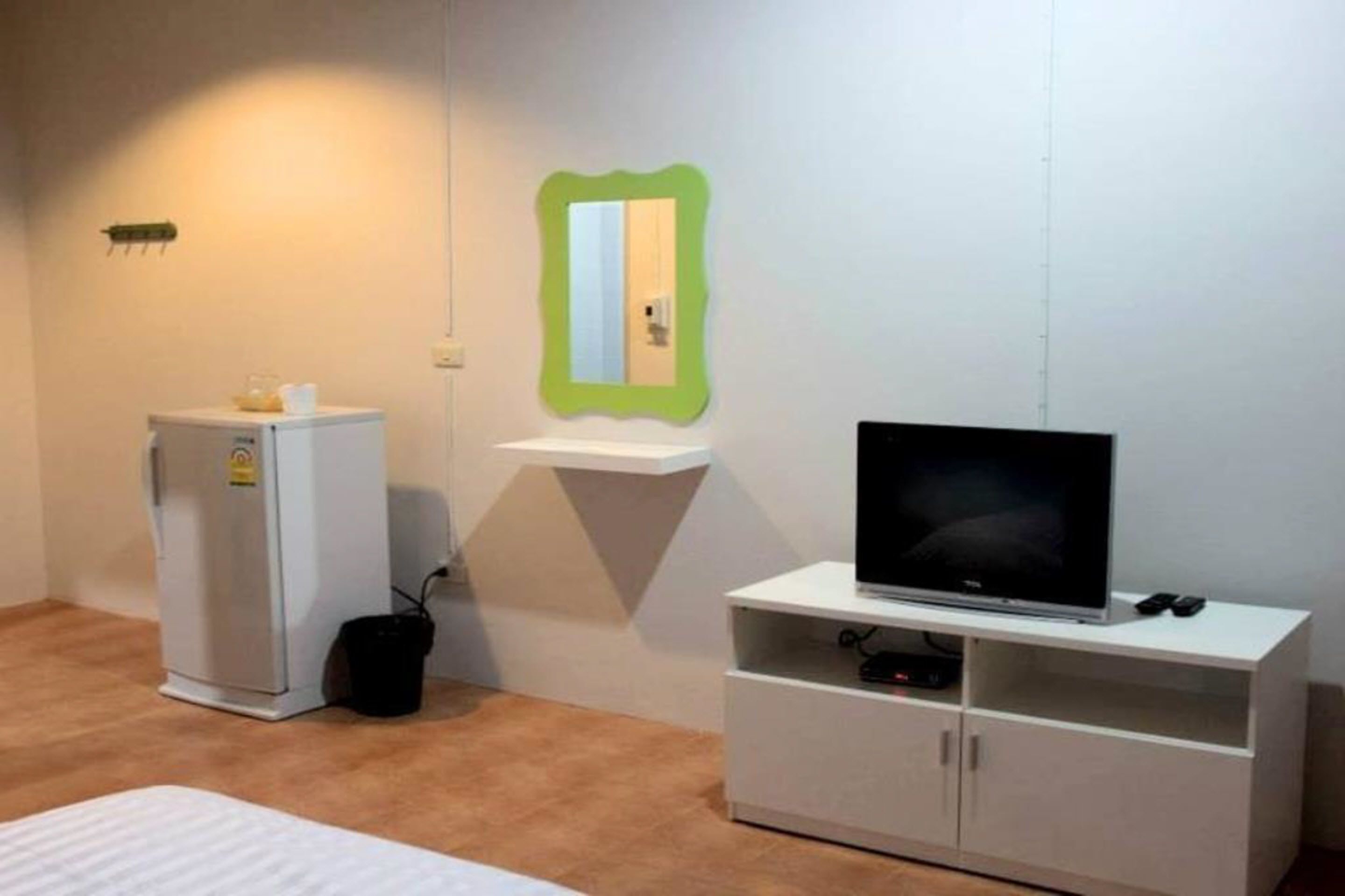 A small room with a bed, a white cabinet with a flat-screen TV, a small refrigerator, and a green-framed mirror on the wall.