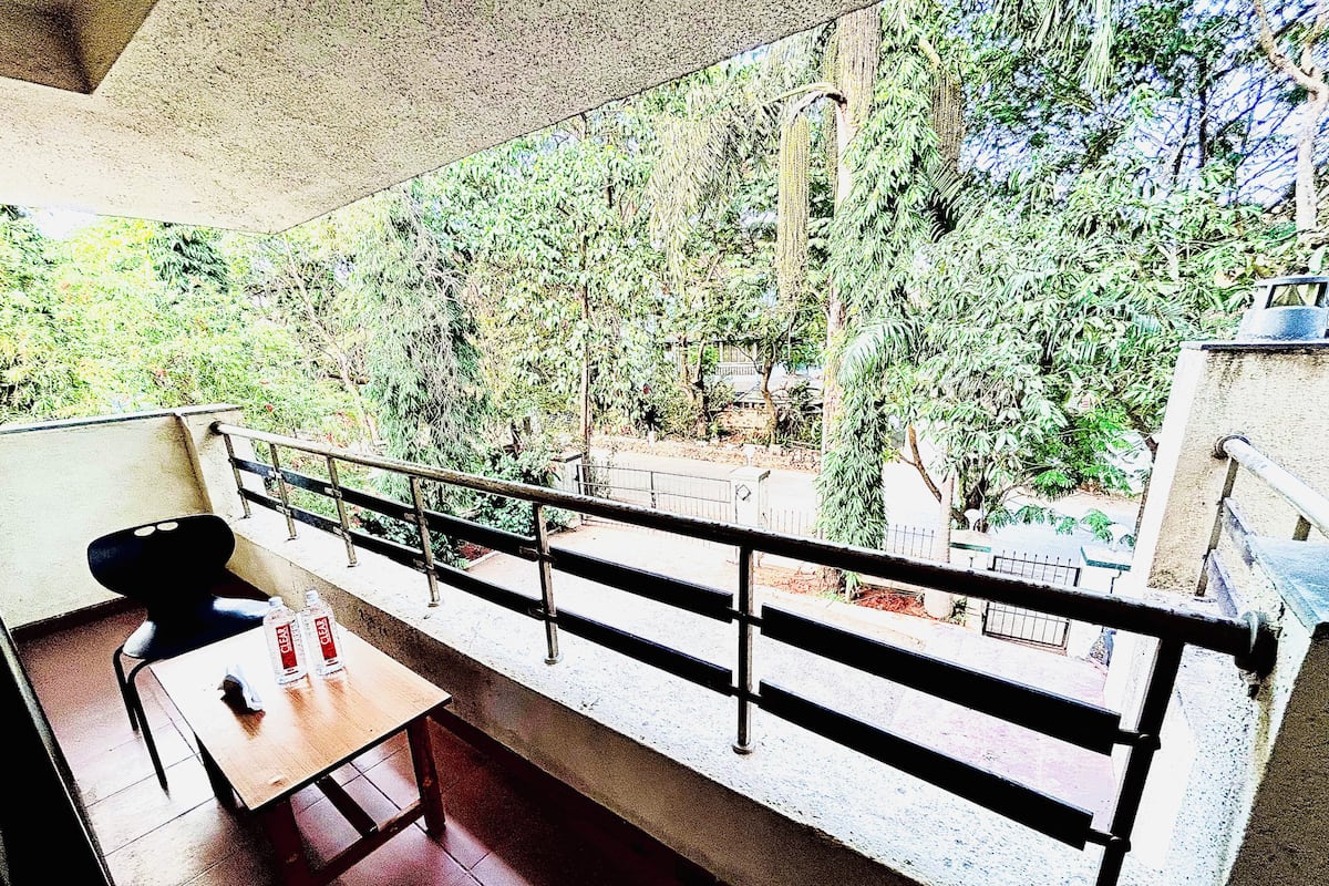Ein Balkon mit Tisch und Stühlen, einem Geländer und Blick auf einen Garten mit Bäumen und einem Gebäude im Hintergrund.
