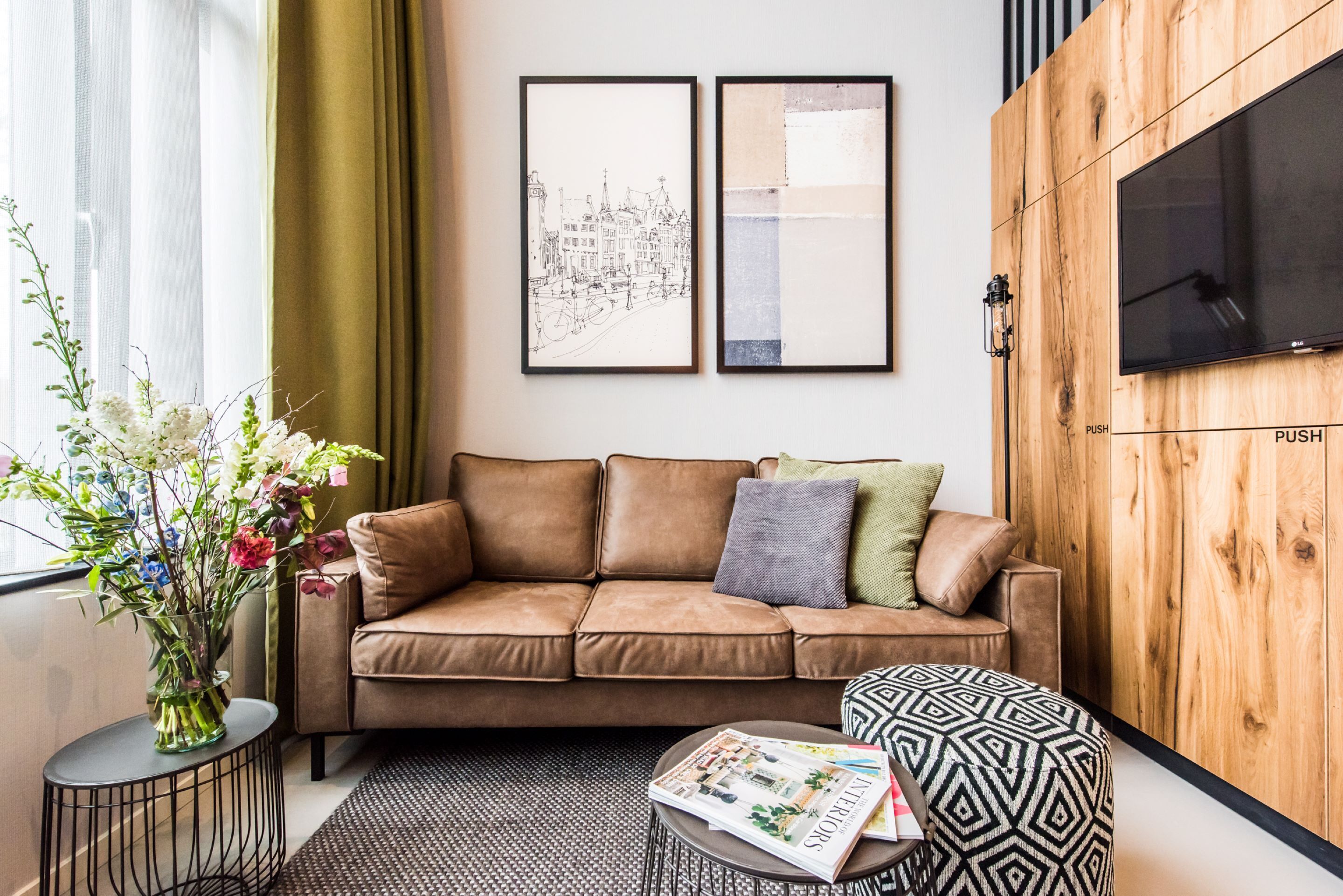 Ein modernes Wohnzimmer mit einem braunen Ledersofa, einem runden Couchtisch mit Zeitschriften, einem gemusterten Hocker, einer Holz-Akzentwand und einem Wand-Fernseher.