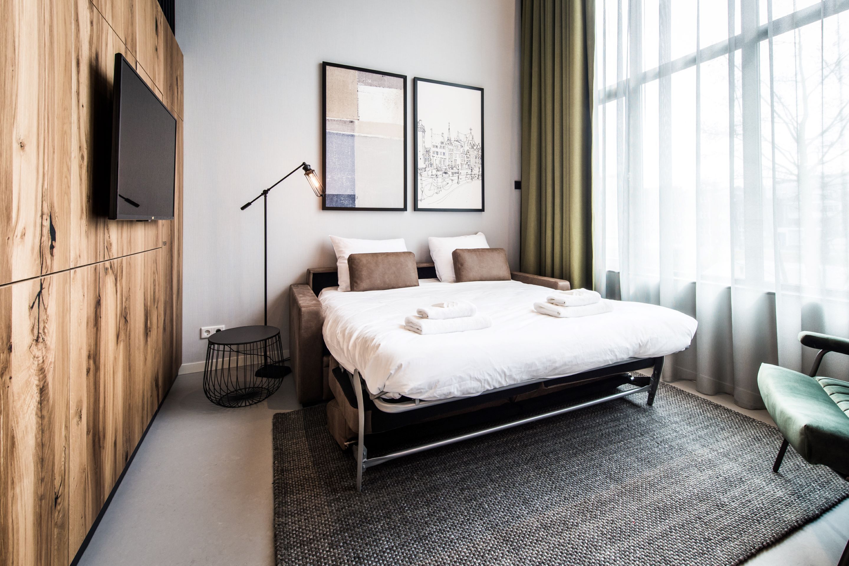 A modern hotel room with a bed, two pillows, a bedside table, a lamp, and a view of a cityscape through the window.