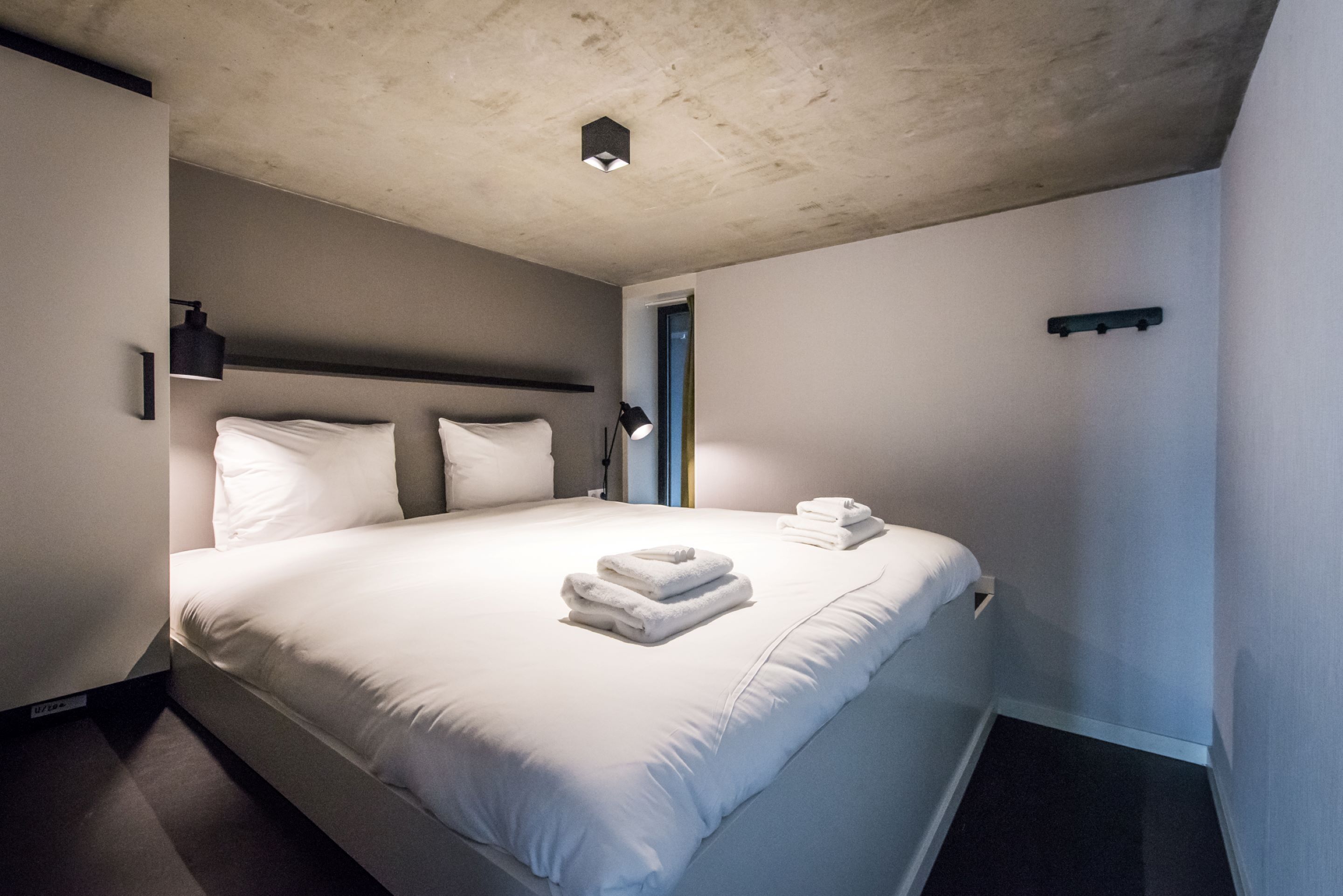 Een hotelkamer met een groot bed, witte lakens, een hoofdbord, een bedlamp en een deur.