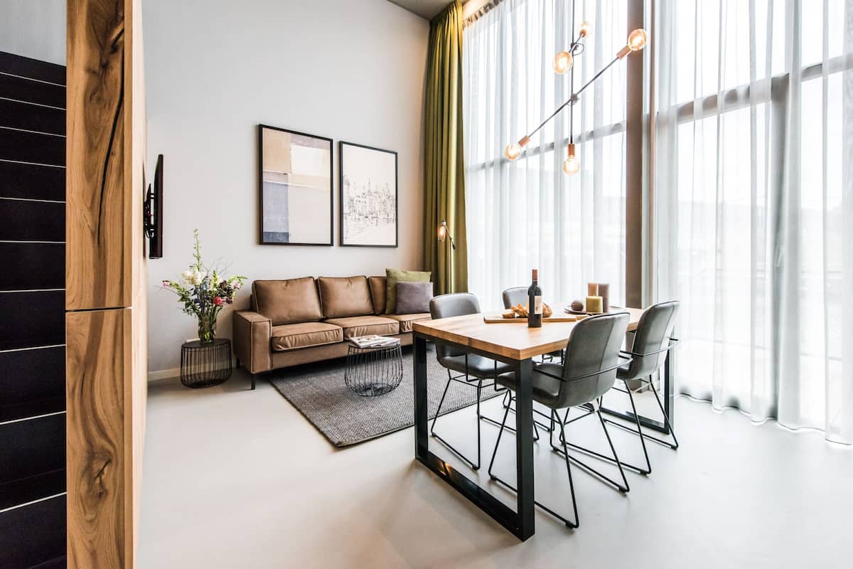 Ein modernes Wohnzimmer mit Holzboden, einer Couch, einem Esstisch mit Stühlen und einem großen Fenster mit transparenten Vorhängen.