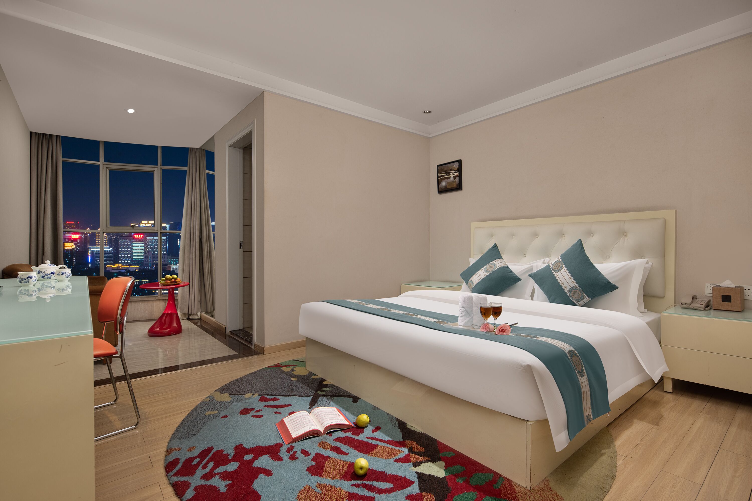 Habitación de hotel con una cama grande, un escritorio con una silla, una mesita con fruta y vistas a la ciudad de noche.