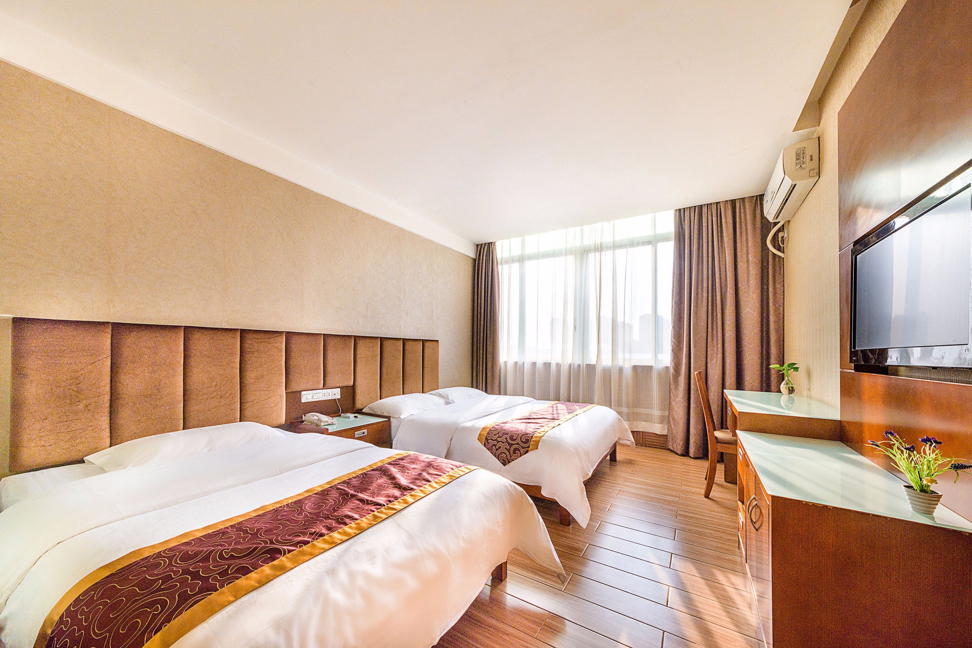 A hotel room with two beds, a desk, a TV, and a window with curtains.