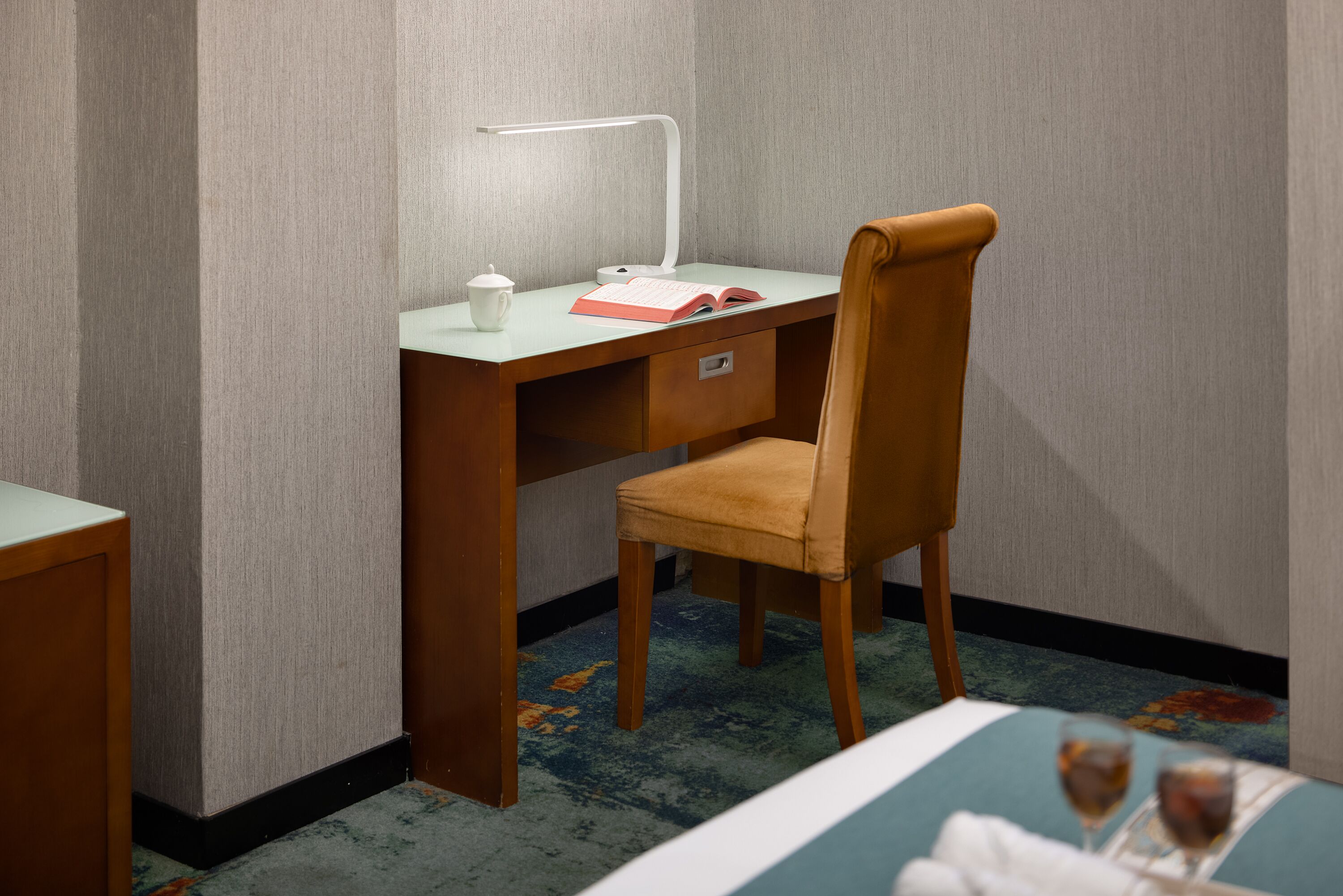Habitación de hotel con escritorio de madera, una silla, una lámpara, un libro y un vaso de agua sobre la mesa.