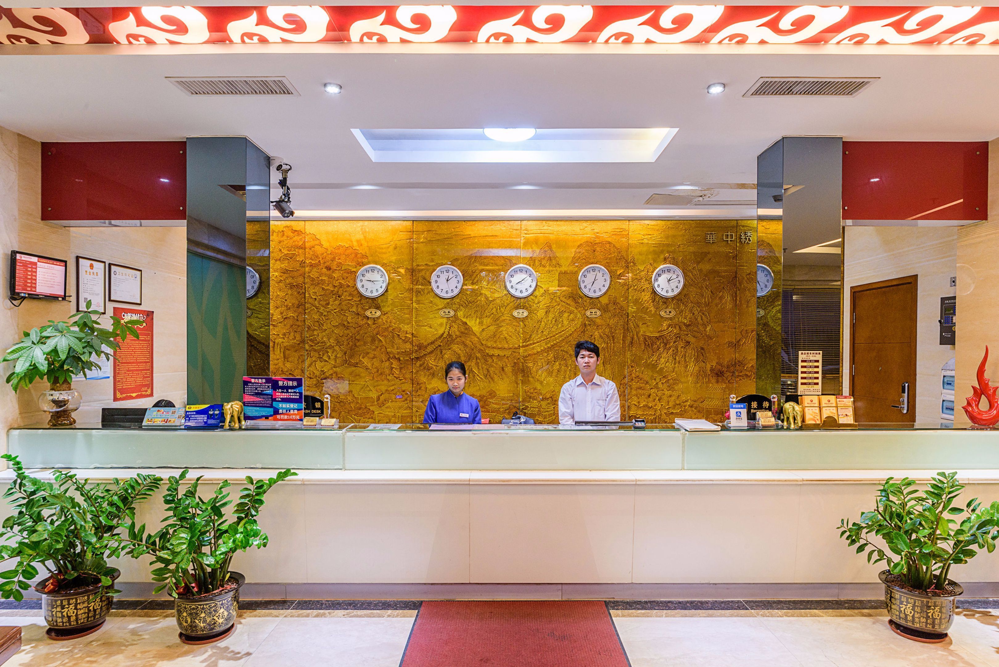 A hotel reception desk with two staff members, a clock wall, and potted plants.