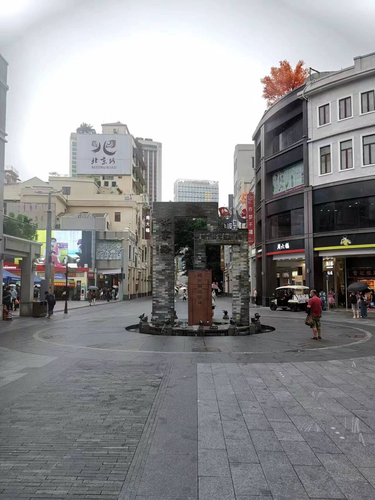 A public square with a central stone monument, surrounded by buildings with commercial establishments.
