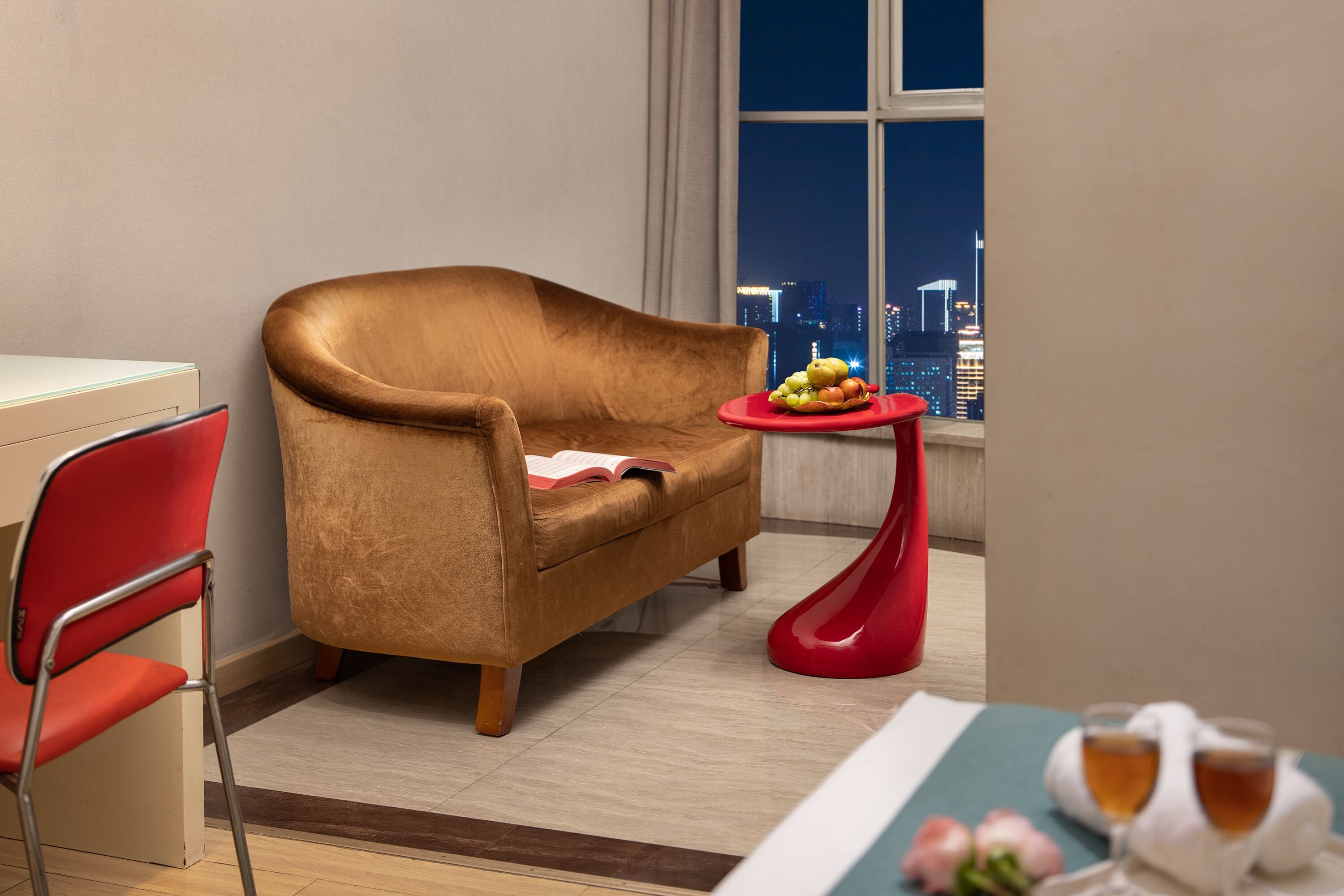 A modern living room with a brown sofa, a red chair, a small red table with fruits, and a view of a cityscape at night.