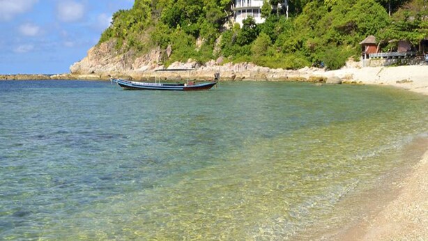 Un bote sobre aguas claras cerca de una playa con un edificio en un acantilado al fondo.