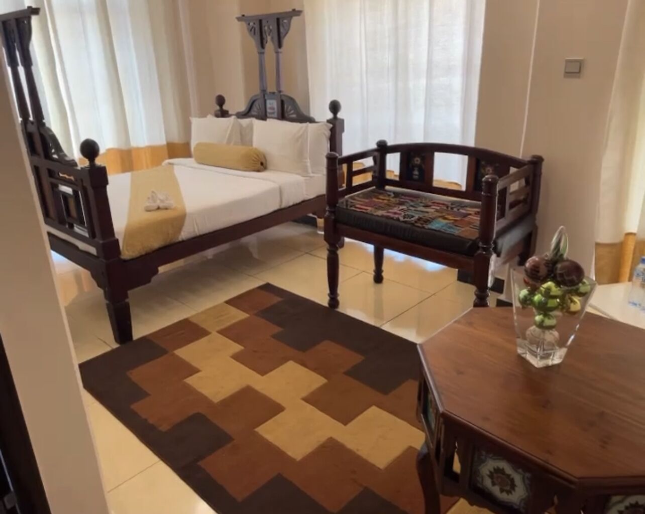 A bedroom with a bed, a wooden chair, a table with a vase, and a patterned rug.