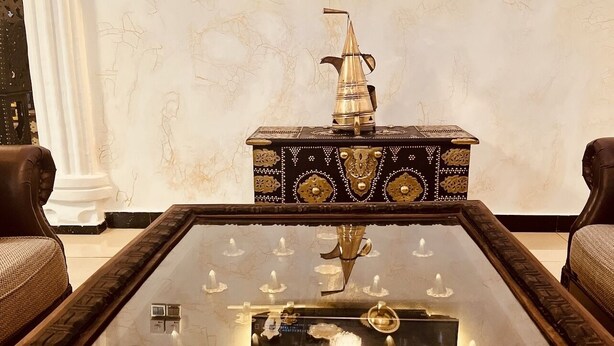 Un salon luxueux, doté d’une grande table basse au plateau réfléchissant, d’un pichet décoratif en laiton et d’un téléviseur fixé au mur, qui diffuse une émission.