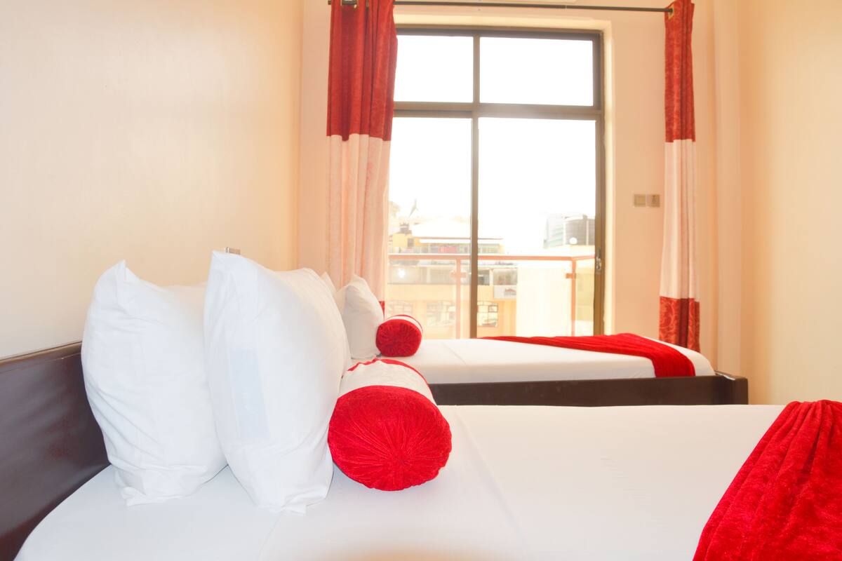 Une chambre d’hôtel avec deux lits, du linge de lit blanc, des touches de couleur rouge et une grande fenêtre avec des rideaux.