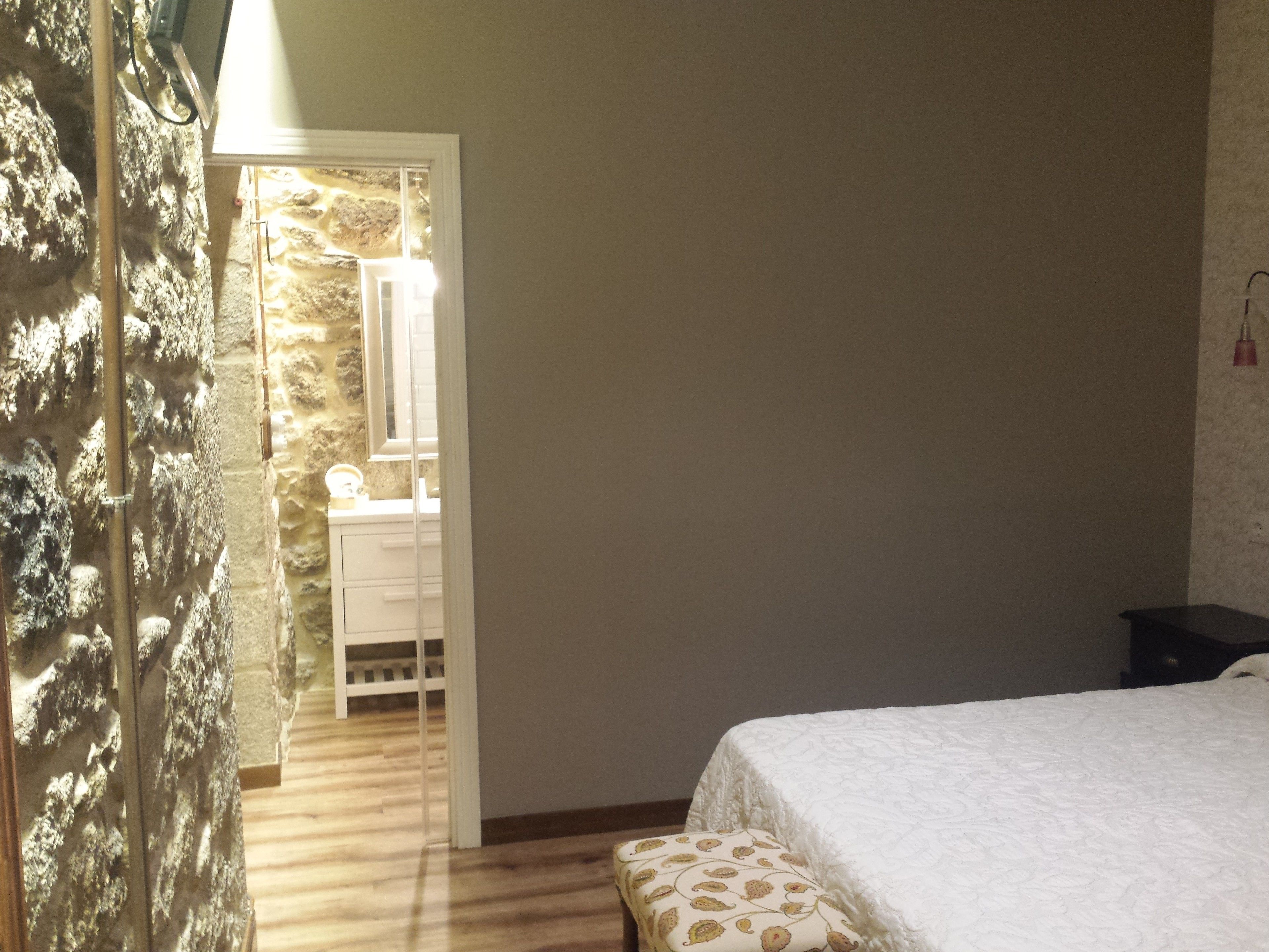 Un dormitorio con cama, piso de madera, pared de piedra y baño con espejo y lavamanos.