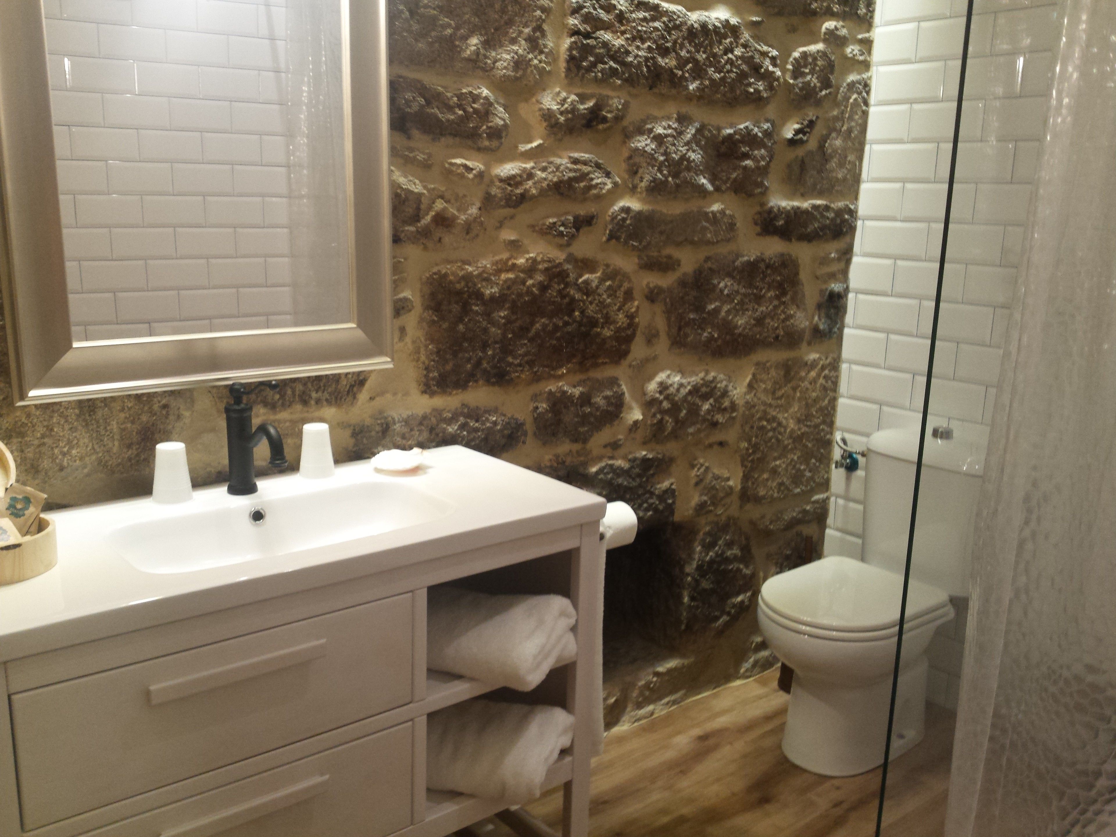 Un baño con lavabo blanco, espejo, pared de piedra, inodoro y zona de ducha.