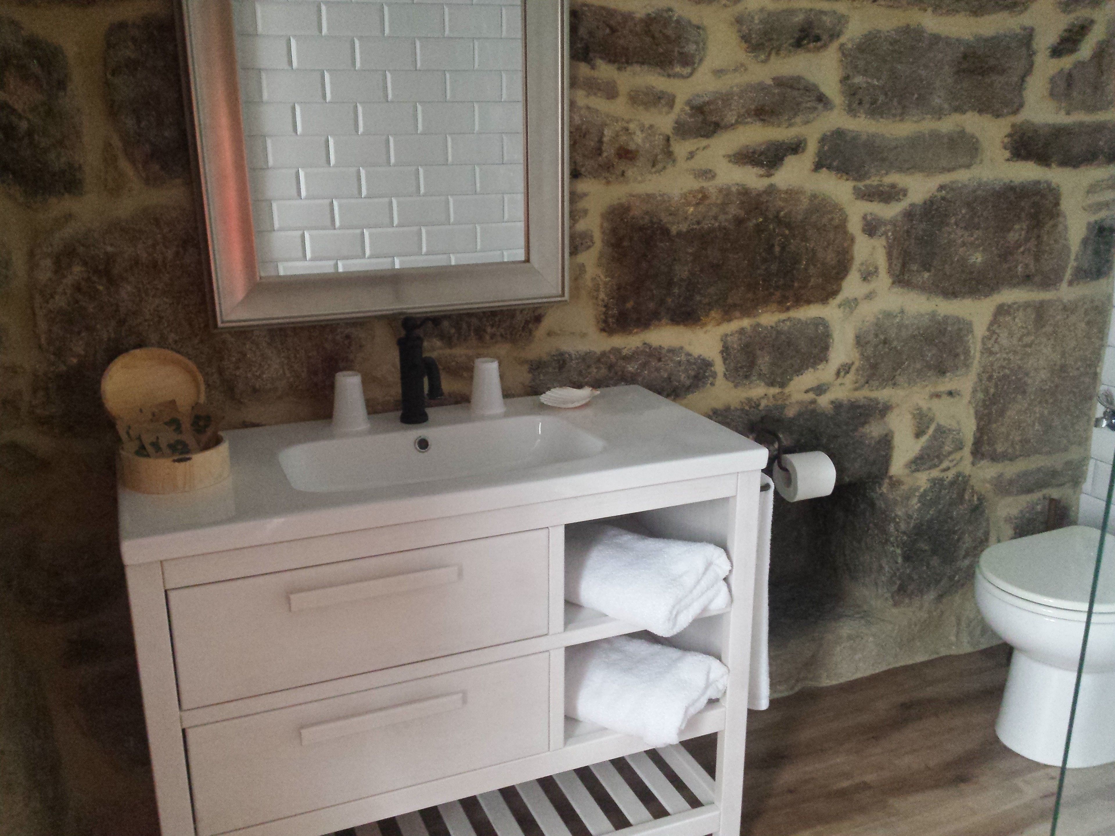 A bathroom with a white sink, a mirror with a brick pattern, a stone wall, a toilet, and a wooden cabinet with towels.