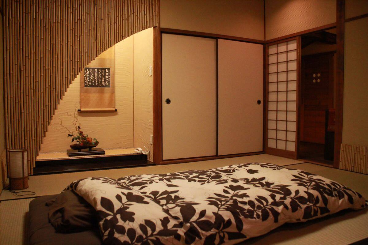 Une pièce de style japonais traditionnel, avec un sol recouvert de tatamis, une petite table ornée d’un arrangement floral, des portes coulissantes et un lit recouvert d’une courtepointe à motifs floraux.