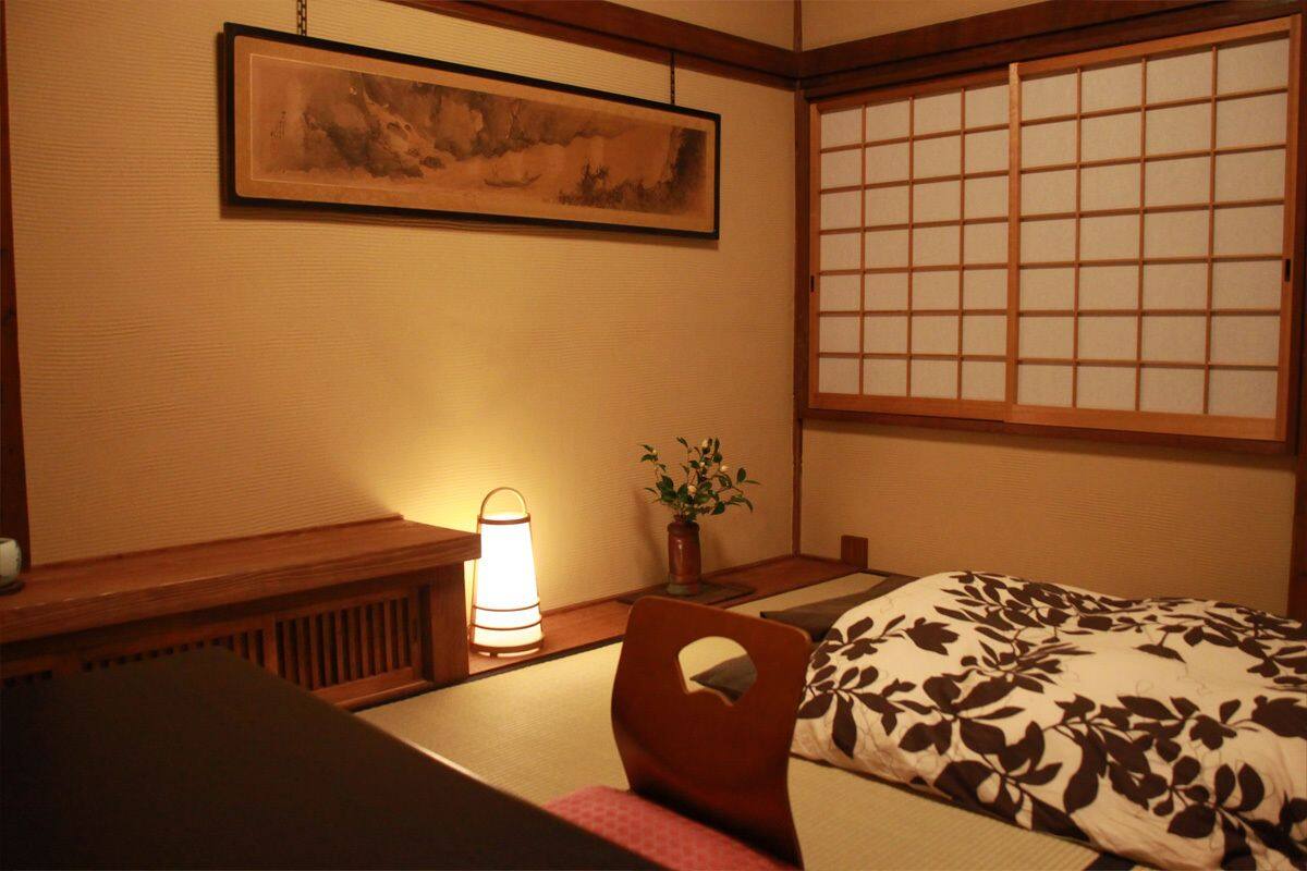 Une pièce de style japonais traditionnel, avec un sol recouvert de tatamis, un petit bureau en bois, un tableau accroché au mur, un paravent coulissant de type shoji et une table de chevet avec une lampe.