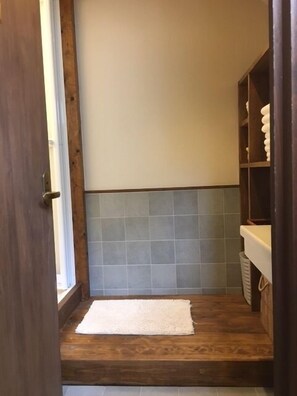 Un baño con suelo de madera, una toalla blanca en el suelo, pared de azulejos y estante de madera con toallas.