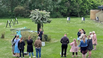 Ulkoilmatapahtuma, jossa ihmiset seisovat ja keskustelevat ruohikkoisella alueella. Taustalla näkyy puita ja taideteoksia.