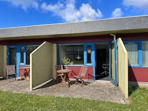 A building with a red exterior, blue-framed windows, and a door. There is a small outdoor seating area with a table and chairs. - Hotel Kobæk Strand (Skælskør)