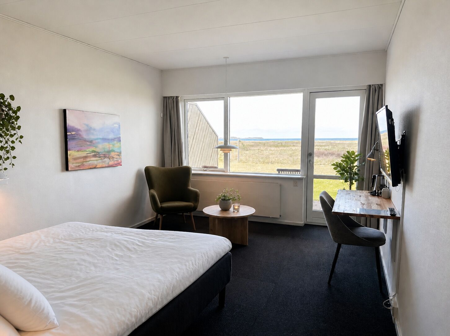Habitación de hotel con cama, escritorio, silla, mesita, planta en maceta, un cuadro y ventana con vistas.