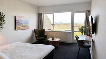 Habitación de hotel con cama, escritorio, silla, mesita, planta en maceta, un cuadro y ventana con vistas.
