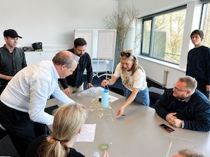 Eine Gruppe von Personen in einem Besprechungsraum mit einer Tafel, einem Fenster mit Blick auf Bäume und einem Tisch mit Papieren und einem Modellgebäude.