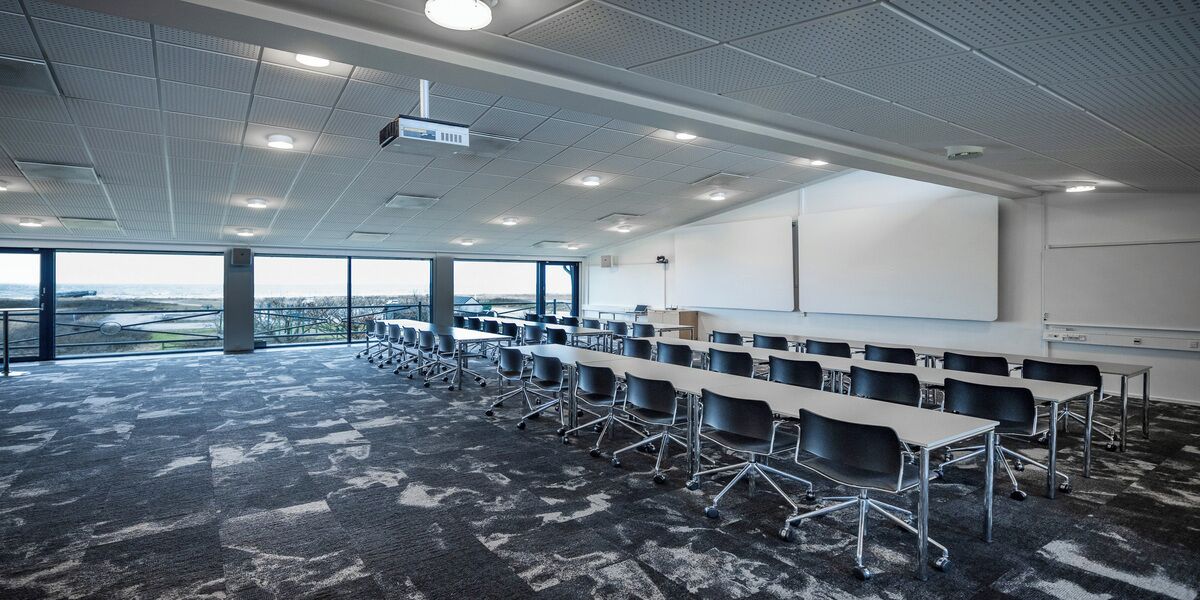 A conference room with rows of tables and chairs, a projector, a whiteboard, and large windows offering a view of the outdoors.