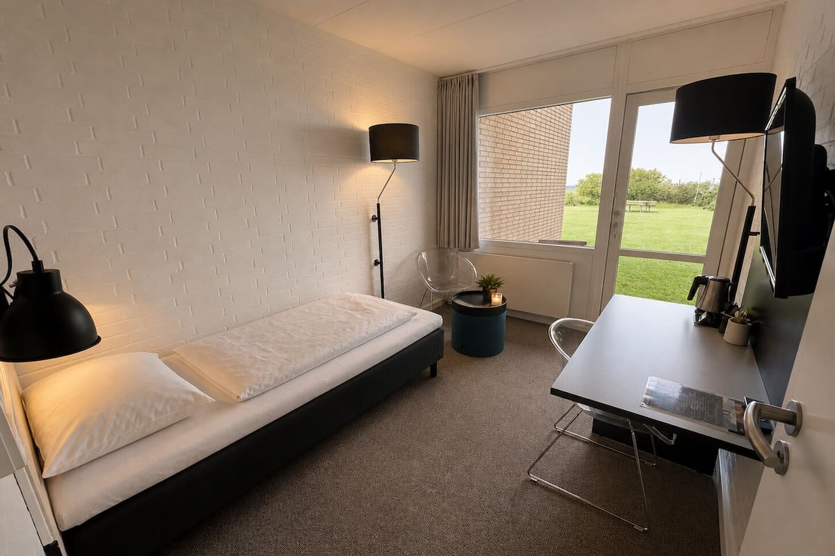 A modern hotel room with a bed, a desk, a chair, a lamp, and a view of the outdoors.