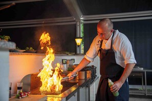 A chef grilling food outdoors with flames, a grill, and cooking utensils. - Hotel Kobæk Strand (Skælskør)