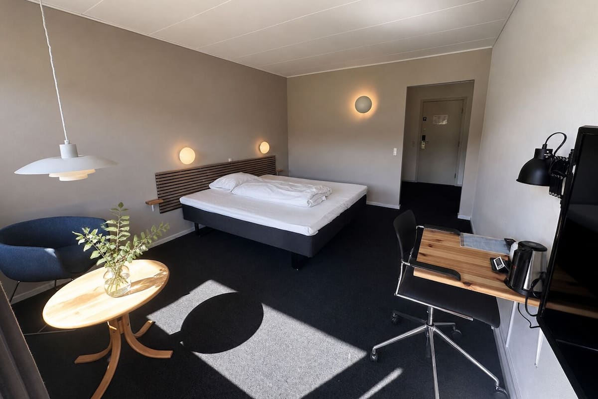 A modern hotel room with a bed, desk, chair, and a small table with a plant.
