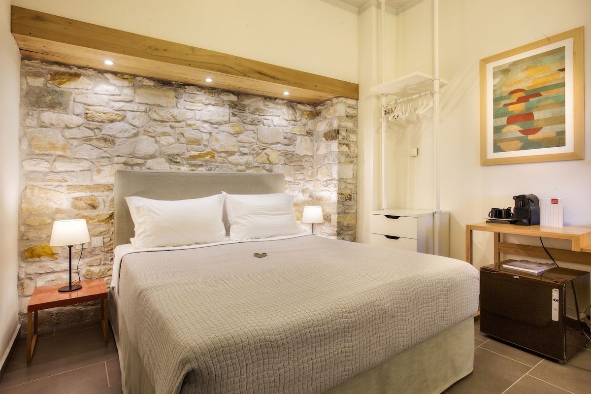 Un dormitorio con pared de piedra, una cama con ropa blanca, una mesita de noche con lámpara, un cabecero de madera, una pequeña mesa con cafetera y un cuadro abstracto enmarcado en la pared.