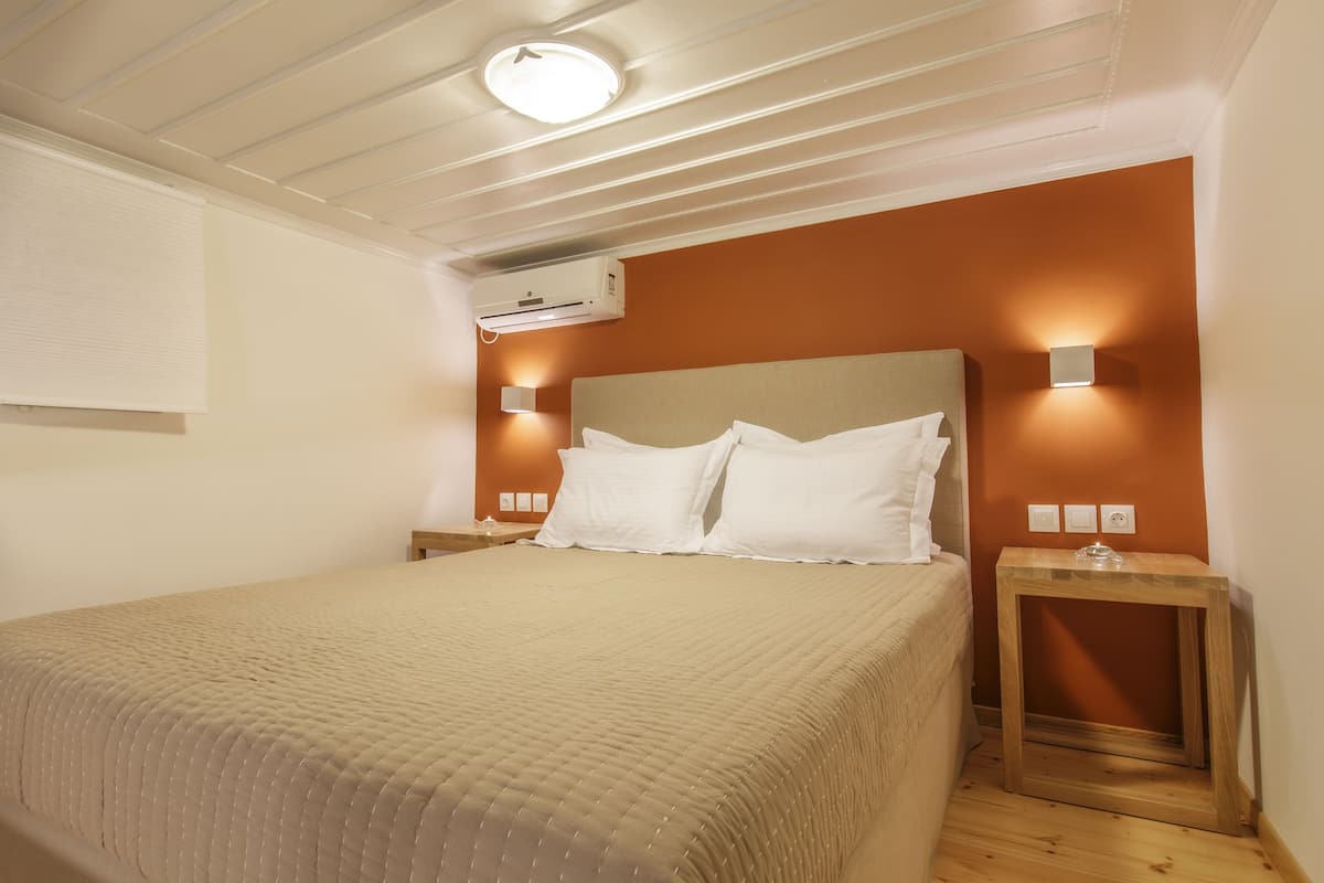 Una cama bien hecha con colcha color beige, una mesita de noche de madera con un objeto de vidrio, y un aire acondicionado instalado en la pared.