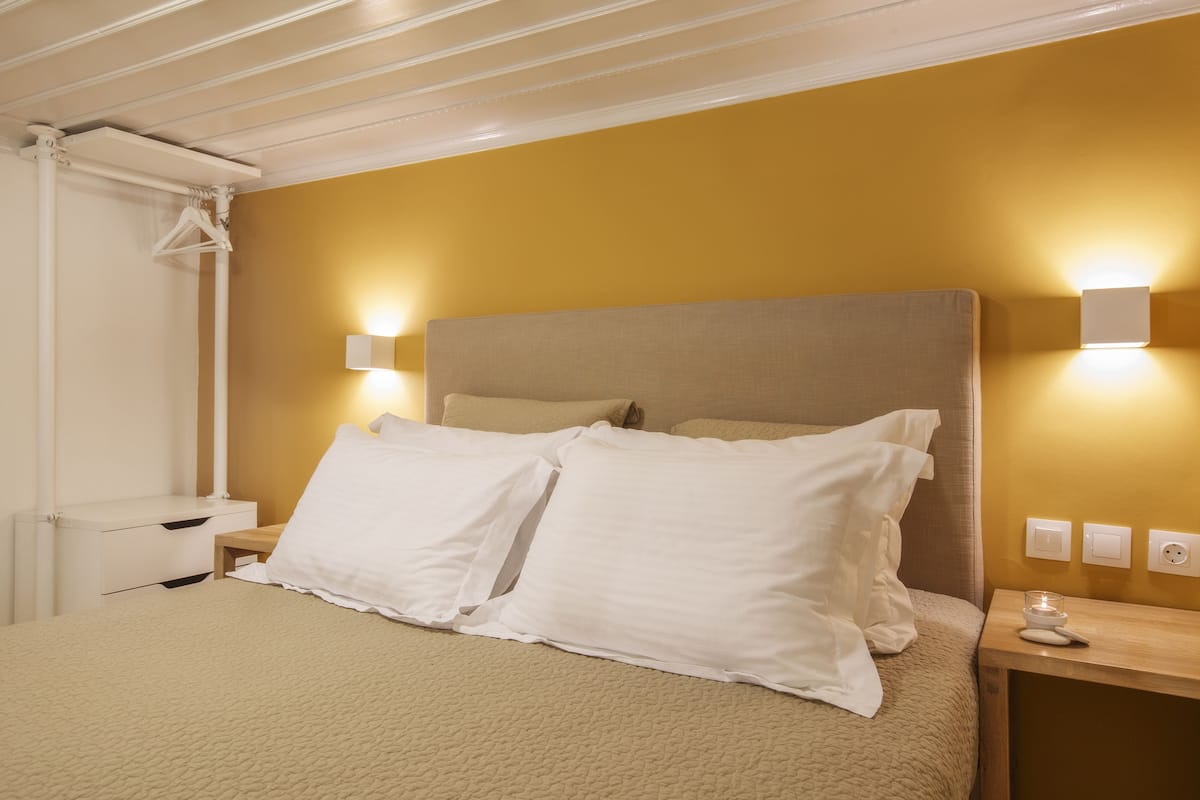 Una cama bien hecha con ropa blanca, una mesita de noche de madera con una lámpara y un aplique en la pared.