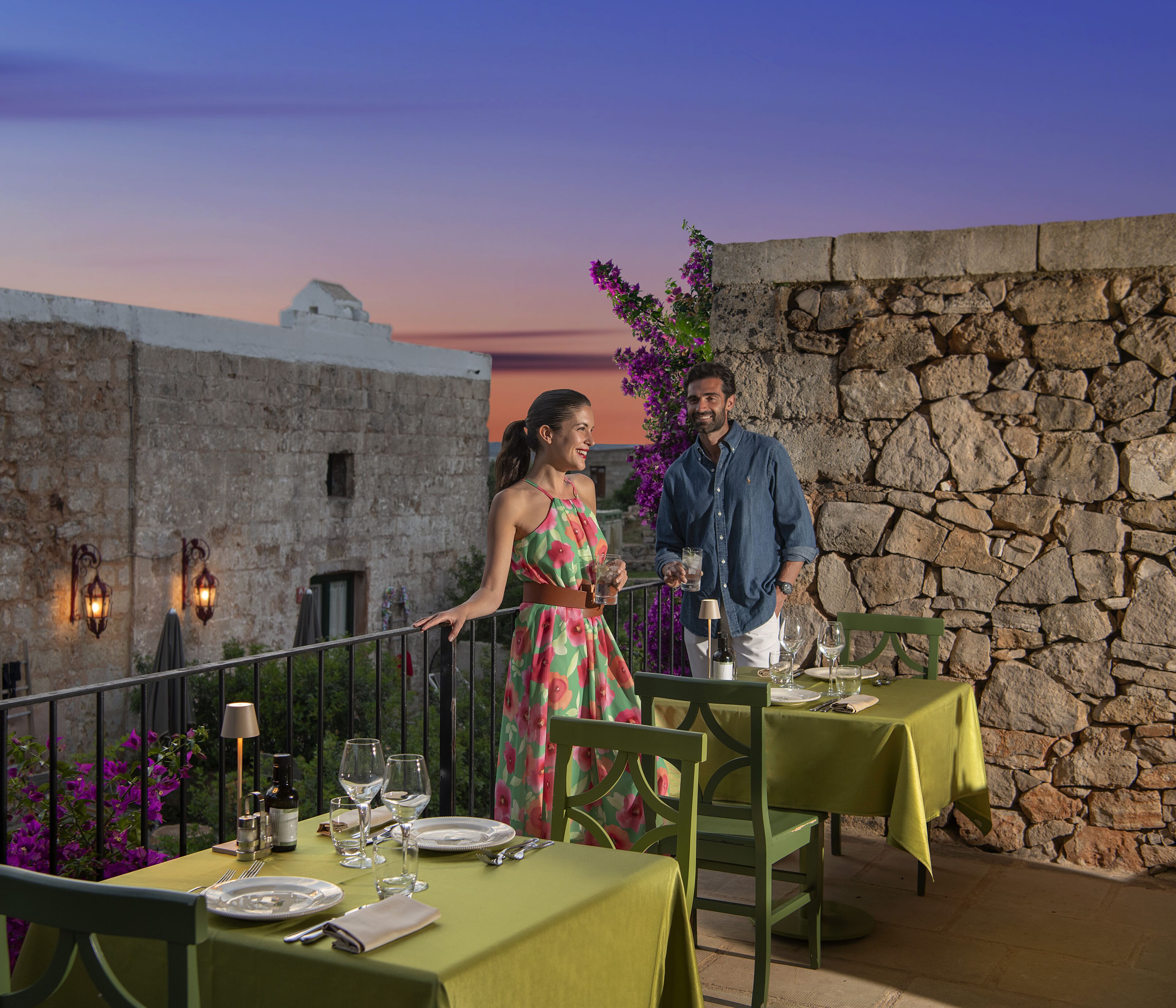 Una terraza con muros de piedra, una mesa lista para cenar y un atardecer de fondo.